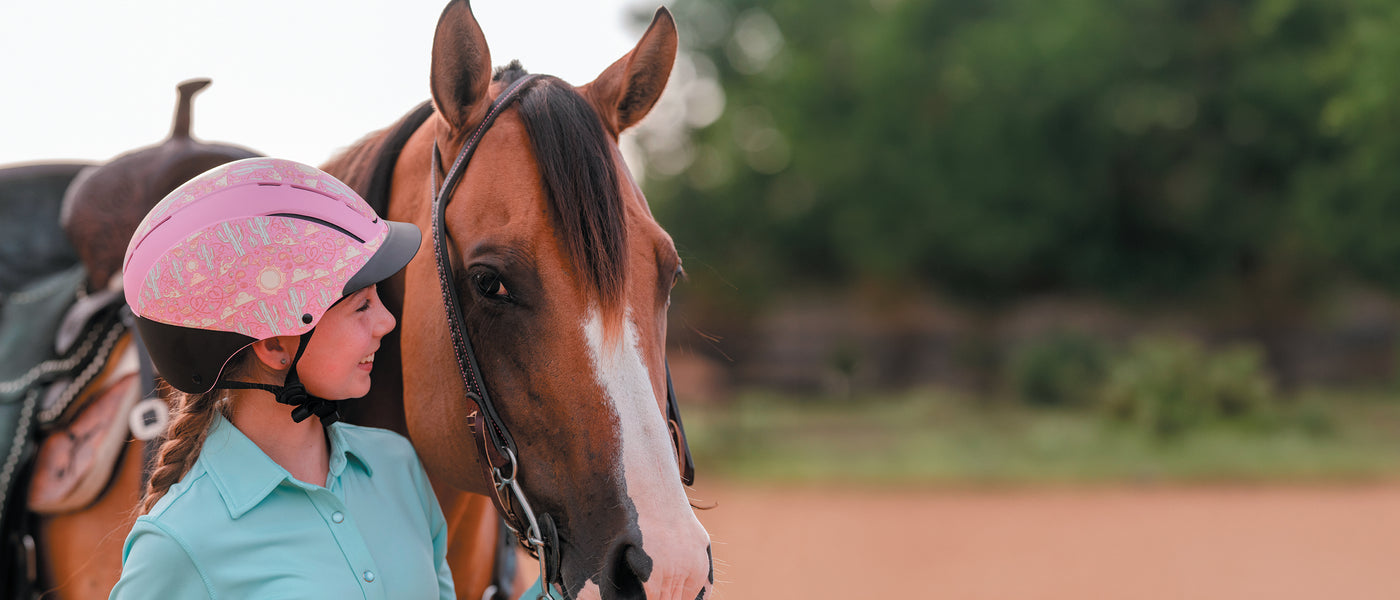 Young rider wearing a pink Troxel helmet beside a brown horse.