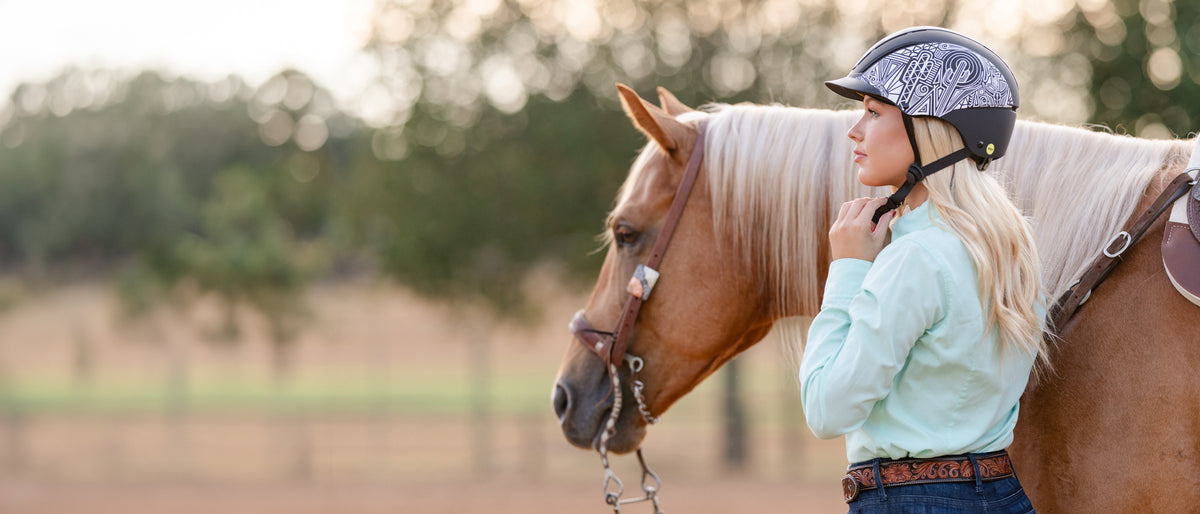 Woman adjusting her riding helmet beside a saddled horse outdoors.