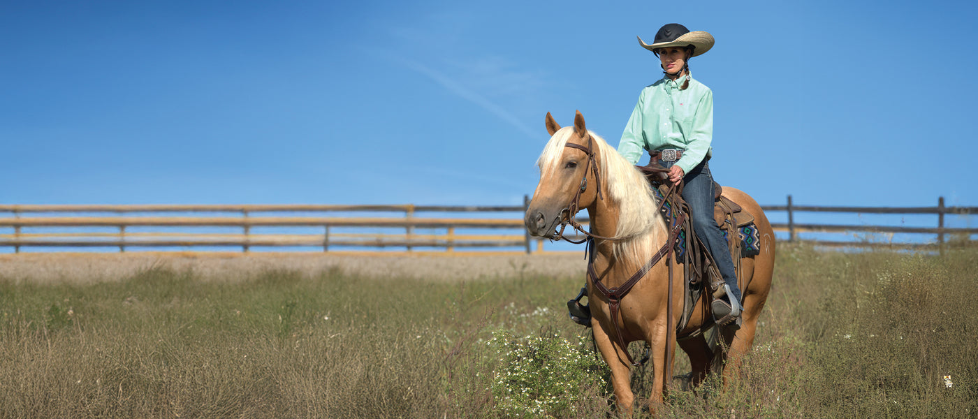 Rider wearing a wide-brim equestrian helmet rides a palomino horse through a grassy pasture under a clear blue sky.