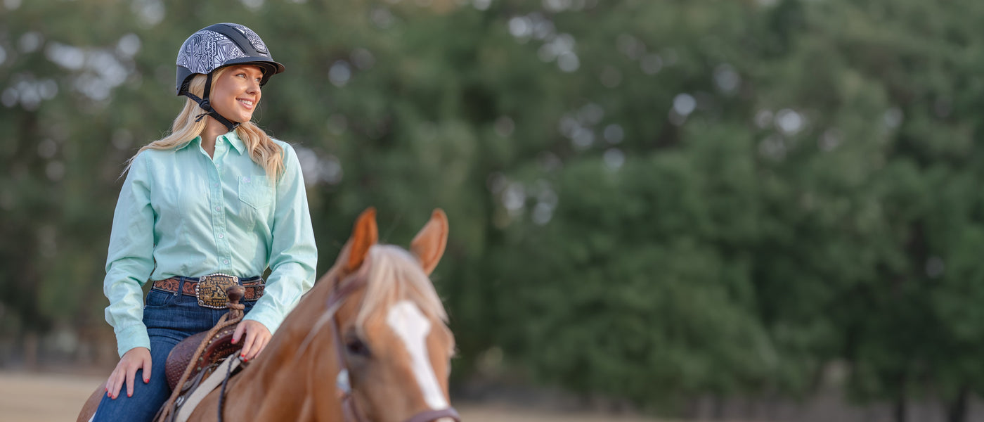 Woman wearing a riding helmet smiles while riding a chestnut horse outdoors.